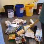 Ein Haufen Müll liegt auf dem Fußboden. An der Wand stehen vier Mülleimer. Je einer für Restmüll, Papier/Pappe, Gelber Sack und Biomüll.