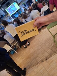 Ein Schwamm mit der Aufschrift KLUB UNIVERSUM. Im Hintergrund findet eine Veranstaltung statt.