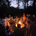 Kinder und Erwachsene stehen und sitzen um eine Feuersschale. An Stöcken haben sie Marshmallows, die sie über das Feuer halten.