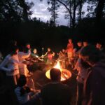 Kinder und Erwachsene stehen und sitzen um eine Feuersschale. An Stöcken haben sie Marshmallows, die sie über das Feuer halten.