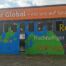 Eingang in eine Halle die mit einer halben Weltkugel bemalt ist. Zudem stehen die Worte Fairtrade, Rohstoffe, Nachhaltiger Leben und Recycling auf der Halle. Oben drüber steht auf einem orangen Schild in weißen Buchstaben "Tour Global - mit uns auf Spurensuche"