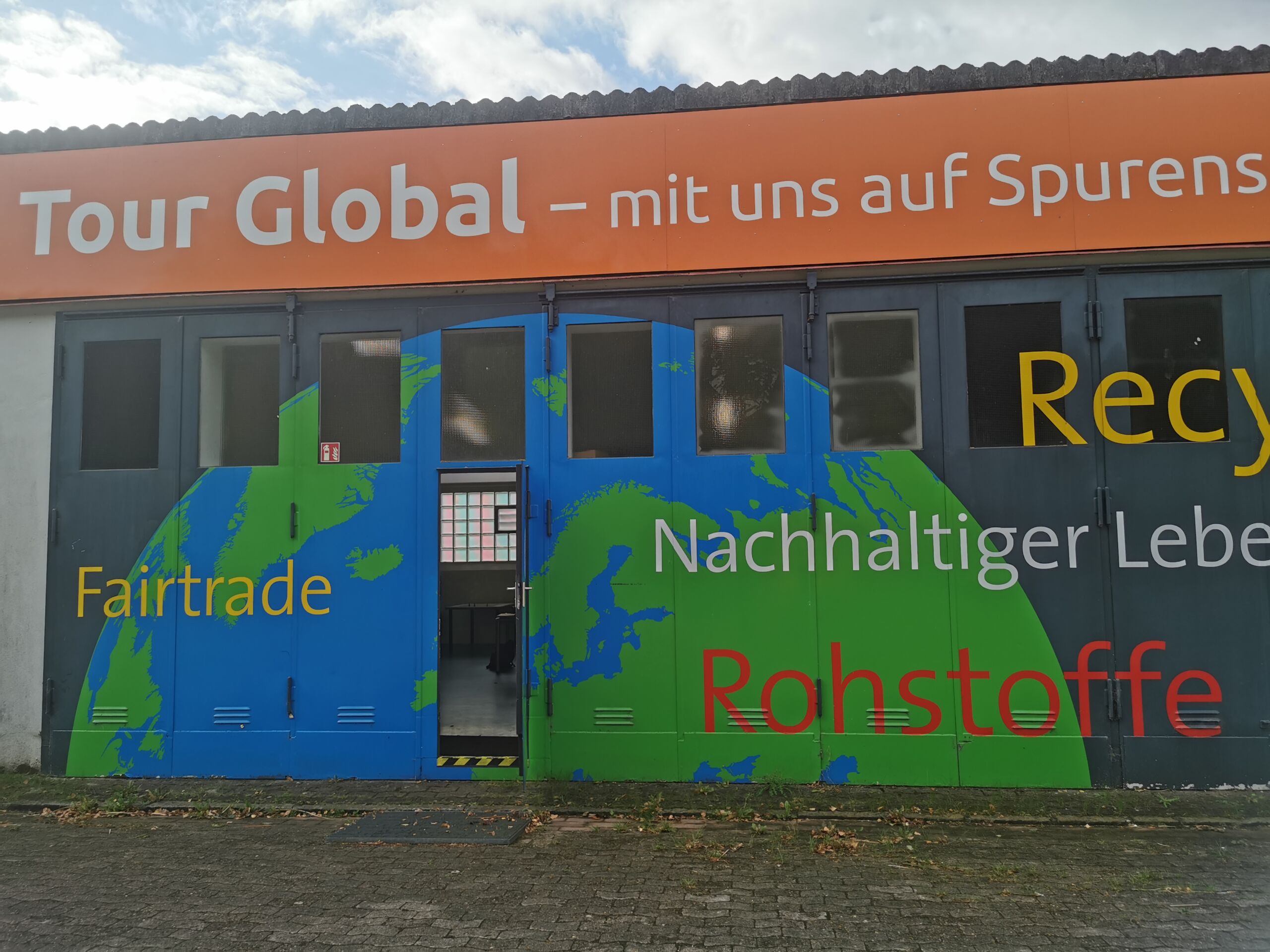 Eingang in eine Halle die mit einer halben Weltkugel bemalt ist. Zudem stehen die Worte Fairtrade, Rohstoffe, Nachhaltiger Leben und Recycling auf der Halle. Oben drüber steht auf einem orangen Schild in weißen Buchstaben "Tour Global - mit uns auf Spurensuche"