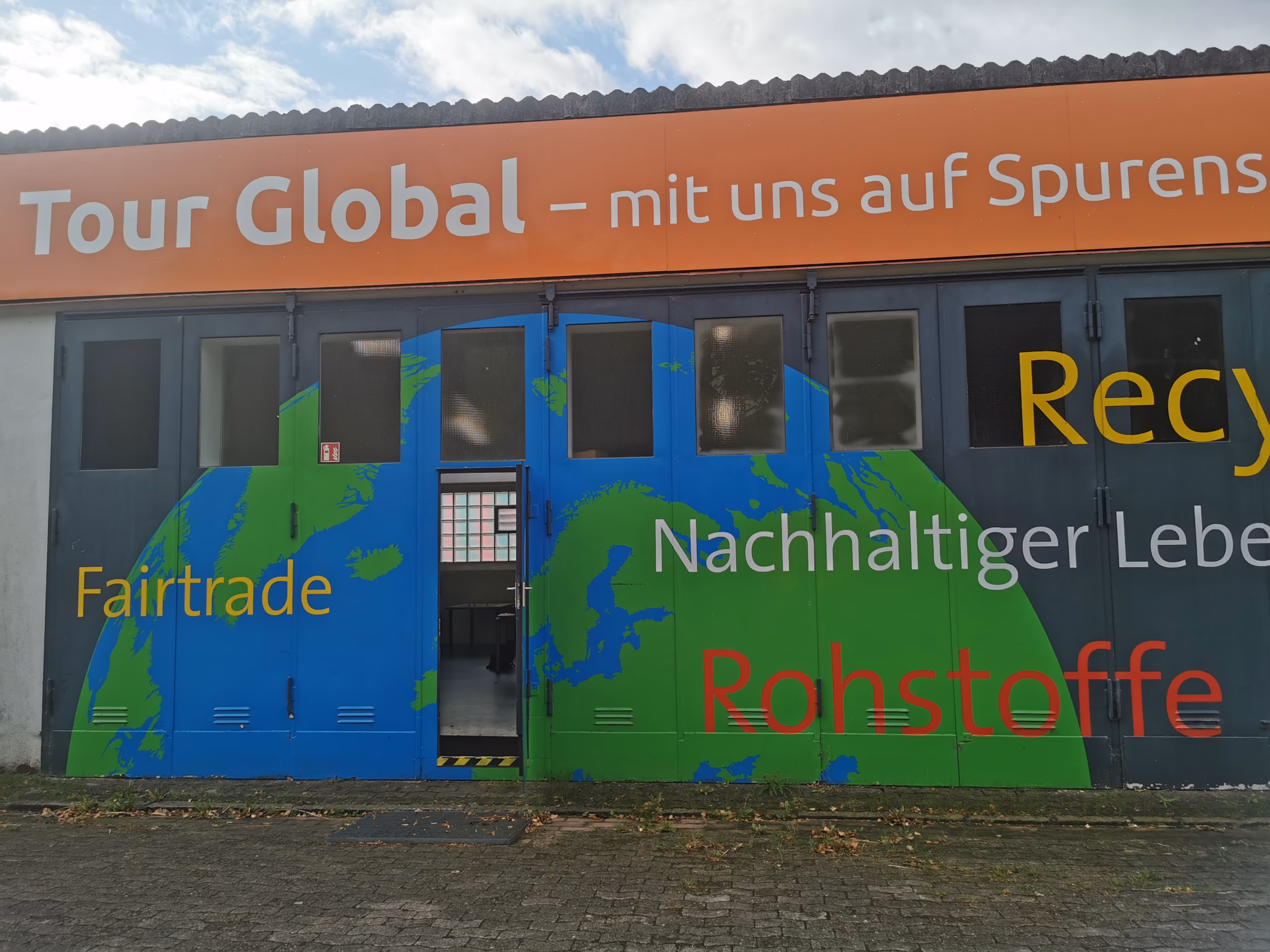 Eingang in eine Halle die mit einer halben Weltkugel bemalt ist. Zudem stehen die Worte Fairtrade, Rohstoffe, Nachhaltiger Leben und Recycling auf der Halle. Oben drüber steht auf einem orangen Schild in weißen Buchstaben "Tour Global - mit uns auf Spurensuche"