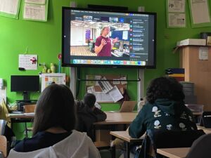 Eine Klasse schaut in einem Klassenraum gespannt auf den großen Bildschirm auf dem eine Liveschalte zu einem Forschungsschiff läuft. Aktuell ist auf dem TV eine Frauen zu sehen, die etwas berichten.