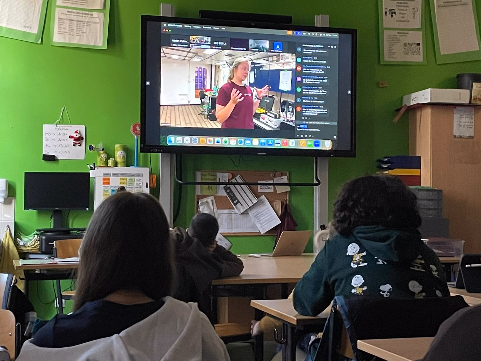 Eine Klasse schaut in einem Klassenraum gespannt auf den großen Bildschirm auf dem eine Liveschalte zu einem Forschungsschiff läuft. Aktuell ist auf dem TV eine Frauen zu sehen, die etwas berichten.
