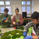 Drei Mädchen kleben Moos mit grünem Kleber in ein rundes, weißes Holzgestell.