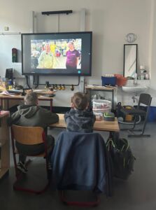 Zwei Schüler schauen in einem Klassenraum gespannt auf den großen Bildschirm auf dem eine Liveschalte zu einem Forschungsschiff läuft. Aktuell sind auf dem TV zwei Frauen zu sehen, die etwas berichten.
