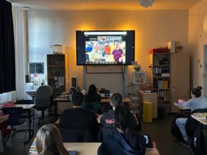 Eine Klasse schaut in einem Klassenraum gespannt auf den großen Bildschirm auf dem eine Liveschalte zu einem Forschungsschiff läuft. Aktuell sind auf dem TV zwei Frauen zu sehen, die etwas berichten.