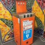 Ein Wasserspender der vor einem bunten Mosaik steht. Der Wasserspender ist mit einer orangen Folie beklebt.