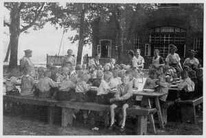 Ein Gruppe Kinder sitzt auf Holzbänken vor dem Schullandheim Ristedt. Begleitpersonen sind ebenfalls zu sehen. Das Bidl ist mehrere Jahrzenhte alt.
