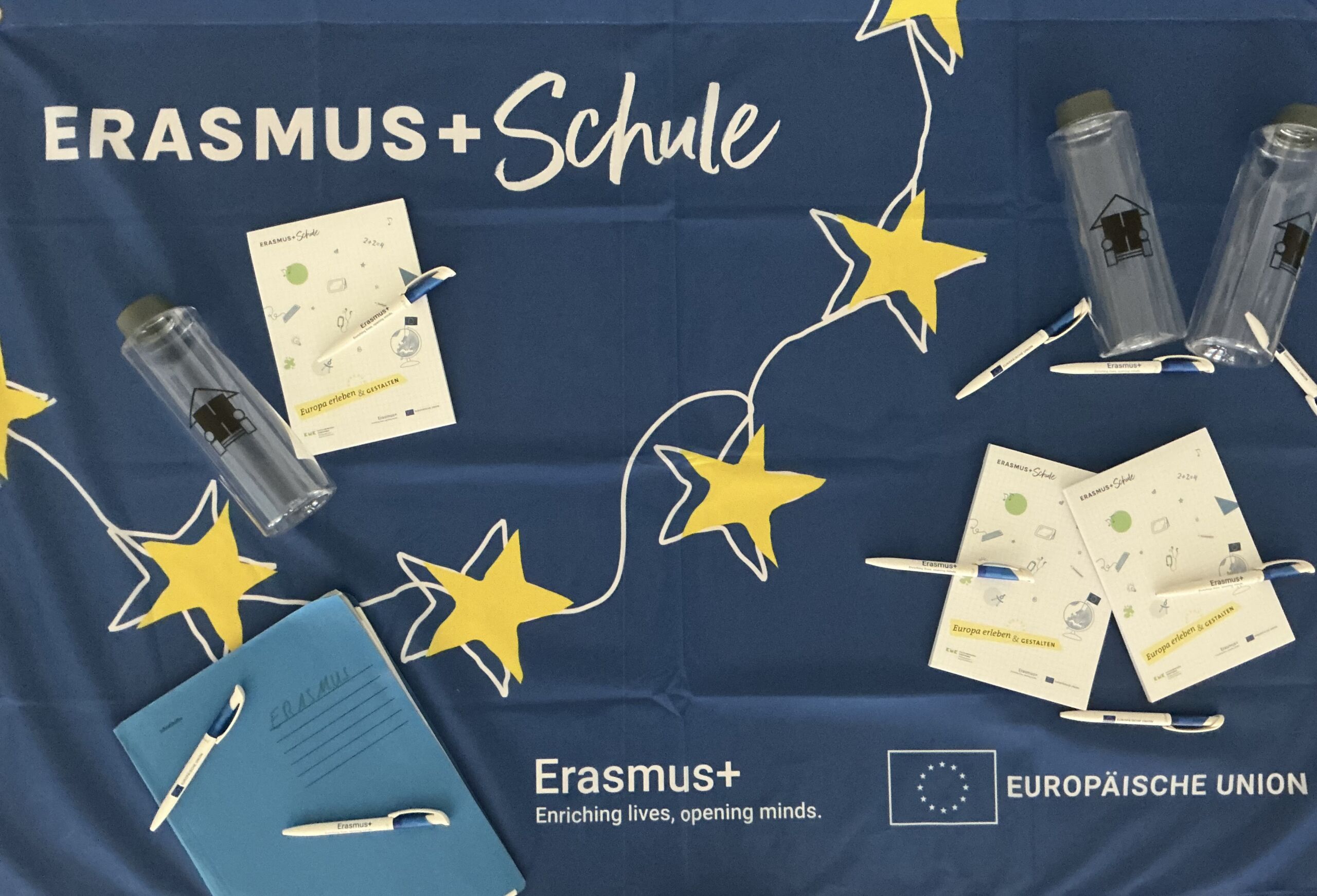 Eine blaue Fahne von Erasmus+ mit mehreren Blöcken, Kugelschreibern und Flaschen die auf der Flagge liegen.