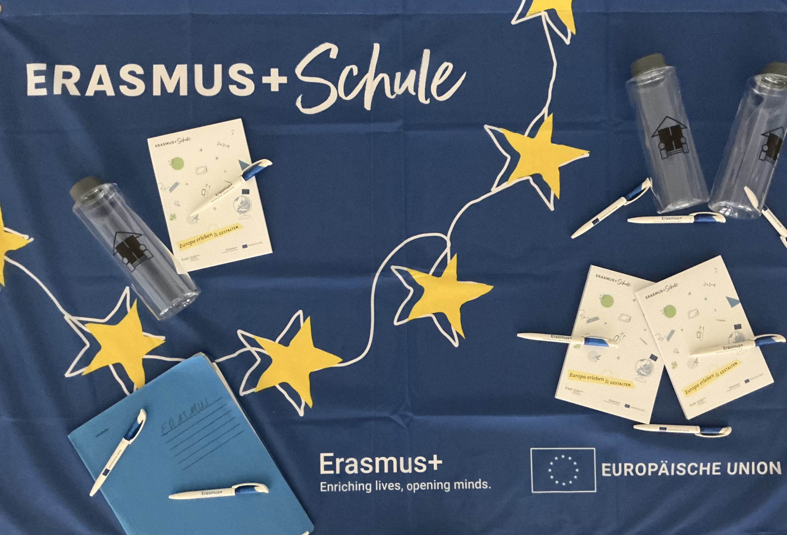 Eine blaue Fahne von Erasmus+ mit mehreren Blöcken, Kugelschreibern und Flaschen die auf der Flagge liegen.