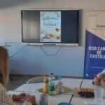 Ein Büro mit zwei Personen die an einem Tisch sitzen und sich unterhalten. Im Hintergrund ist eine Aufsteller von Erasmus+ auf Spanisch zu sehen.