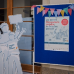 Ein Aufsteller Energiesparmeister und eine Stellwand mit einem Poster mit der Aufschrift "Die besten Klimaschutzprojekte an Schulen gesucht" Dadrüber hängen zwei Wimpel