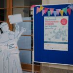 Ein Aufsteller Energiesparmeister und eine Stellwand mit einem Poster mit der Aufschrift "Die besten Klimaschutzprojekte an Schulen gesucht" Dadrüber hängen zwei Wimpel