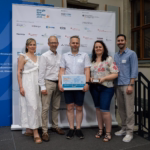 Fünf Personen stehen vor einem Aufsteller mit Werbung. Die Person in der Mittel hält die Plakette "Die besten Klimaschutzprojekte an Schulen" für die Oberschule an der Helgolander Straße Bremen