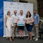 Fünf Personen stehen vor einem Aufsteller mit Werbung. Die Person in der Mittel hält die Plakette "Die besten Klimaschutzprojekte an Schulen" für die Oberschule an der Helgolander Straße Bremen