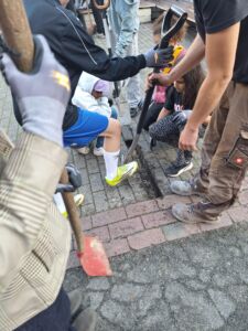 Mehrere Personen, darunter Kinder und Erwachsene, graben mit Schaufeln in einem gepflasterten Boden und haben vier Steine entfernt