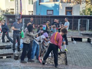 Eine Gruppe von Menschen arbeitet gemeinsam im Freien an einer Bodenfläche, einige halten Werkzeuge und graben Steine aus