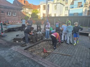 Gruppe von Kindern und Erwachsenen, die auf einem gepflasterten Hof Steine aufnehmen und auf Paletten stapeln, umgeben von Gebäuden und Bauzäunen