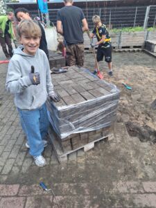 unge mit grauem Kapuzenpullover und blauen Jeans steht lächelnd neben einer Palette mit verpackten Steinen, im Hintergrund weitere Kinder beim Arbeiten im Freien auf sandigem Boden.