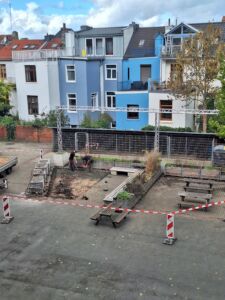 Baustelle mit zwei Arbeitern, die in einem abgesperrten Bereich auf einem gepflasterten Hof arbeiten