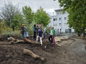 Eine Gruppe von Jugendlichen steht auf einem unbefestigten Gelände mit Baumstämmen und Büschen im Hintergrund