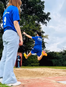 Jugendlicher in blauer Sportkleidung springt beim Sportfest in eine Sandgrube, im Vordergrund steht eine Person mit grauer Jogginghose und blauem T-Shirt mit der Nummer 9