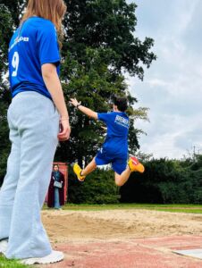Jugendlicher in blauer Sportkleidung springt beim Sportfest in eine Sandgrube, im Vordergrund steht eine Person mit grauer Jogginghose und blauem T-Shirt mit der Nummer 9