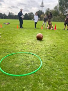 Gruppe von Kindern und einem Erwachsenen auf einer Wiese beim Sportfest mit einem Ball und einem grünen Reifen auf dem Boden