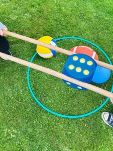 Zwei Personen halten lange Holzstäbe, mit denen sie einen großen blauen Würfel und mehrere Bälle in einem grünen Reifen auf Gras bewegen