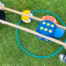 Zwei Personen halten lange Holzstäbe, mit denen sie einen großen blauen Würfel und mehrere Bälle in einem grünen Reifen auf Gras bewegen