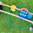 Zwei Personen halten lange Holzstäbe, mit denen sie einen großen blauen Würfel und mehrere Bälle in einem grünen Reifen auf Gras bewegen