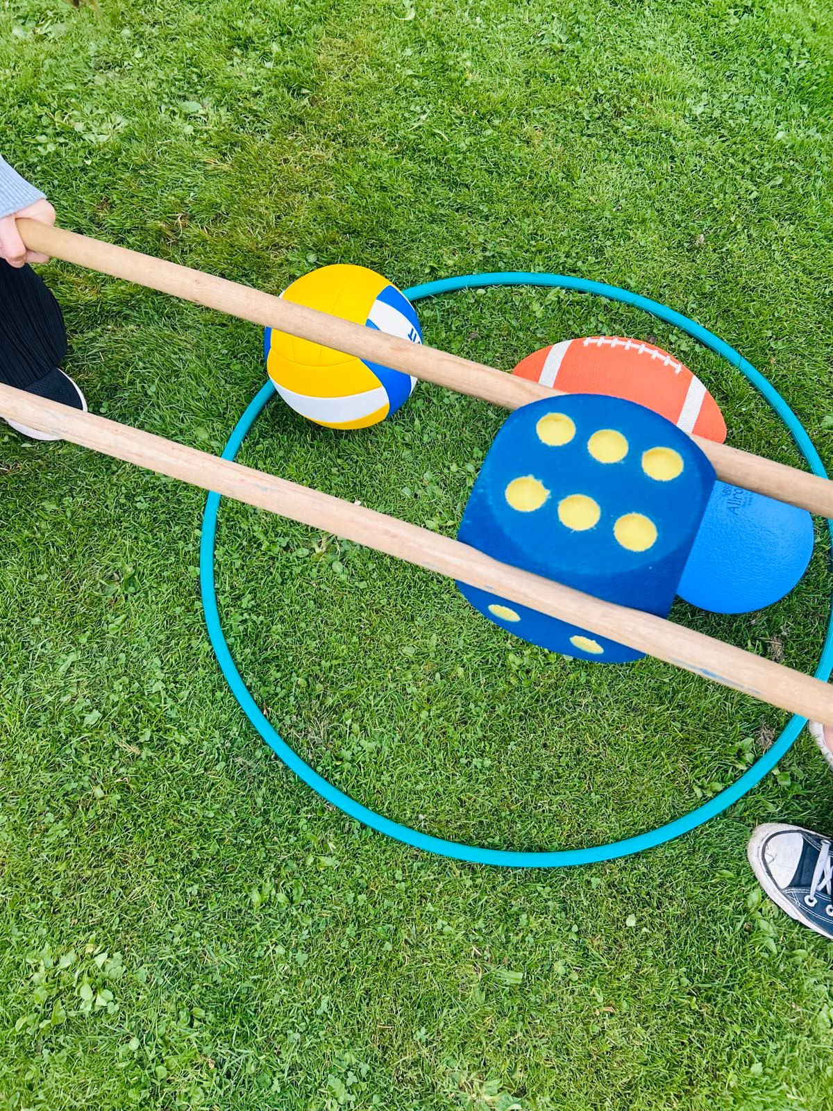 Zwei Personen halten lange Holzstäbe, mit denen sie einen großen blauen Würfel und mehrere Bälle in einem grünen Reifen auf Gras bewegen