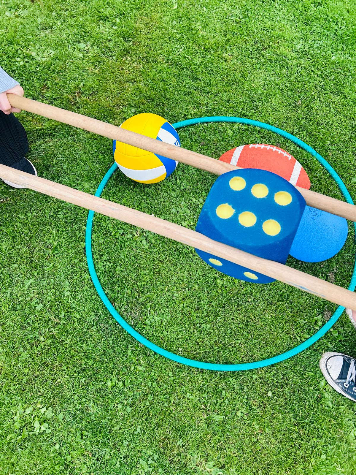 Zwei Personen halten lange Holzstäbe, mit denen sie einen großen blauen Würfel und mehrere Bälle in einem grünen Reifen auf Gras bewegen